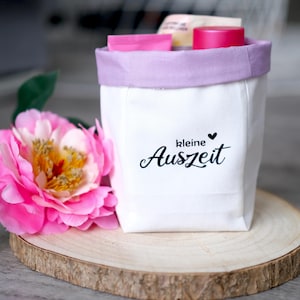 May include: White canvas bag with a purple trim and the text "kleine Auszeit" with a small heart. The bag is filled with pink and white toiletries and is sitting on a wooden slice.