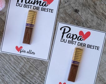 May include: Two white cards with black text and red hearts. One says "Mama DU BIST DIE BESTE" and the other says "Papa DU BIST DER BESTE". Each card has a chocolate bar and the text "für alles".
