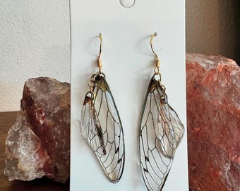 May include: A pair of gold earrings with clear cicada wings. The wings are delicate and translucent, with intricate veins.