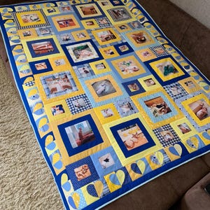 Op de afbeelding: Een handgemaakte quilt met een patchwork-ontwerp met talrijke vierkante fotoblokken. De quilt toont een levendig kleurenpalet van blauw, geel en wit, met een rand van blauwe en gele hartvormen. De quilt wordt getoond op een bruine bank.