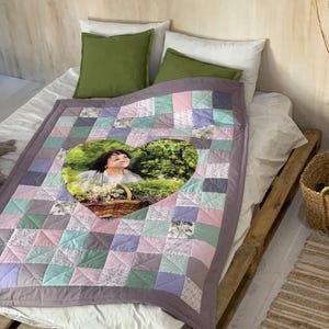 Könnte beinhalten: Eine Patchwork-Decke mit einem herzförmigen Bild einer Person, die einen Korb mit Blumen hält. Die Decke hat Quadrate aus rosa, blauen und grauen Stoffen mit einem lavendelfarbenen Rand. Zwei grüne Kissen und zwei weiße Kissen liegen auf dem Bett.