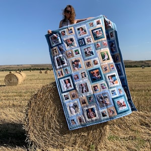 May include: A patchwork quilt with numerous framed photographs, in shades of blue and beige. The quilt is displayed outdoors against a backdrop of a field and a clear blue sky. The quilt is a handmade item.