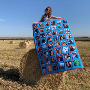 Op de afbeelding: Een blauwe quilt met een rode rand, met talrijke vierkante fotoprints. De quilt wordt omhooggehouden in een veld met hooibalen onder een heldere blauwe lucht. De foto's lijken familieportretten te zijn.
