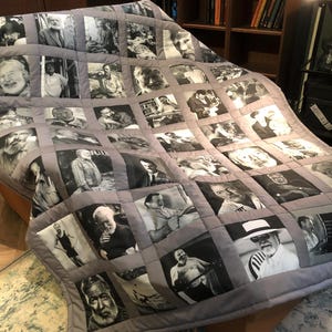 May include: A gray quilt featuring a grid of black and white portraits of various people. The quilt has a gray border and is draped over a bed. The portraits are of different sizes and styles.