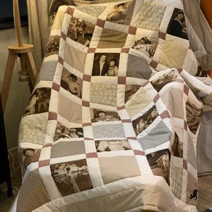 Peut inclure: Une courtepointe faite à la main avec un motif patchwork, présentant des photographies en sépia. La courtepointe incorpore des carrés de différents tissus dans des tons de blanc, beige et marron, avec une bordure mauve. La courtepointe est drapée sur une chaise.