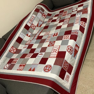 Op de afbeelding: Een handgemaakte patchwork quilt in rood, grijs en wit. Het ontwerp toont geometrische patronen en vormen, waaronder vierkanten, cirkels en gestileerde symbolen. De rand is effen rood met een witte en grijze binnenrand.