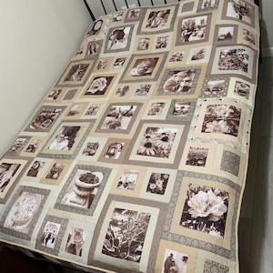 May include: A sepia-toned quilt featuring framed photographs and floral images. The quilt is composed of squares in shades of beige, tan, and brown, with a decorative border. The quilt is placed on a black metal bed frame.