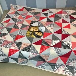 Op de afbeelding: Een handgemaakte quilt met een geometrisch patroon van rode, grijze, zwarte en witte driehoeken. De quilt heeft een centrale foto van twee personen. De driehoeken zijn gemaakt van verschillende stoffen, waaronder bloemen- en polkadotpatronen.