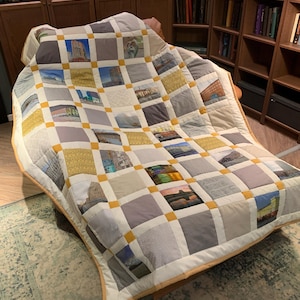 Könnte beinhalten: Eine handgefertigte Patchwork-Decke mit quadratischen Feldern in verschiedenen Farben und Mustern, darunter Bilder von Gebäuden und Landschaften. Die Quadrate sind von weißem Stoff umrandet und mit gelben Akzenten verbunden. Die Decke liegt über einem Stuhl.