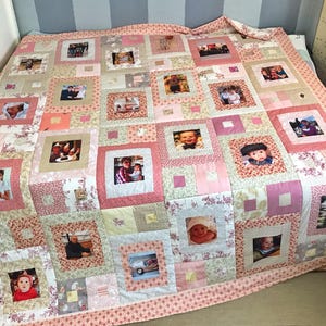 Op de afbeelding: Een handgemaakte patchwork quilt met familiefoto's in vierkante vakken, omgeven door roze en bloemige stoffen. De quilt heeft een roze bloemenrand en ligt op een bed. Het ontwerp is een mix van patronen en kleuren.