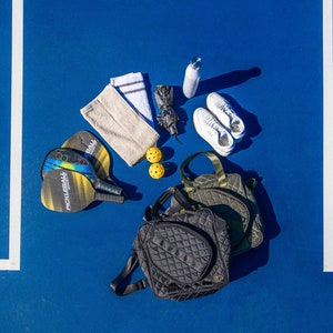 Puede incluir: Una bolsa acolchada negra y verde, dos palas de pickleball negras y amarillas, una toalla blanca, dos pelotas de pickleball amarillas, una botella de agua blanca y dos zapatillas blancas sobre una superficie azul.