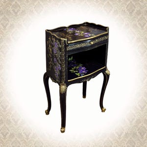Peut inclure: Une table d'appoint en bois noir avec des accents sculptés peints en or sur les pieds et la façade du tiroir. Le dessus et l'étagère intérieure sont décorés de fleurs de clématites violettes et de vignes vertes peintes à la main.