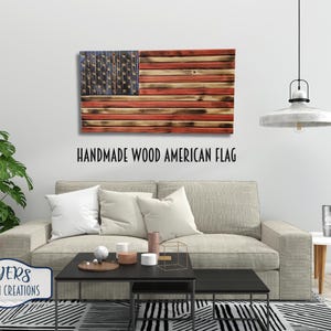 May include: Handmade wooden American flag with red, white, and blue stripes and stars. The flag is hanging on a white wall above a couch and coffee table. The text "HANDMADE WOOD AMERICAN FLAG" is below the flag.