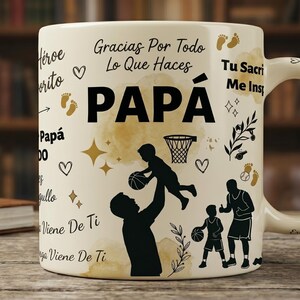 May include: A cream-colored ceramic mug featuring black silhouettes of a parent and child playing basketball. The text reads PAPÁ, Gracias Por Todo Lo Que Haces, and Tu Sacrificio Me Inspira, accompanied by gold watercolor accents, hearts, stars, and footprints.