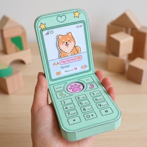 May include: A mint green paper craft flip phone featuring a screen with a cartoon dog illustration and text bubbles that read Hey furrend!, Nyaa!, and Miss U!. The keypad includes a central pink paw print button and standard numeric keys.