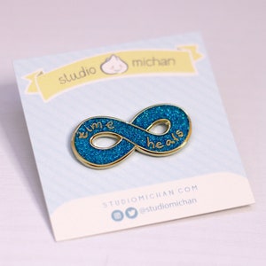 Peut inclure: Épinglette en forme d'infini dorée avec des paillettes bleues et les mots "time heals" inscrits dessus.