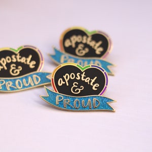 May include: A gold heart-shaped pin with the text "apostate & proud" in black lettering on a black background. The heart is surrounded by a blue ribbon with rainbow glitter accents.