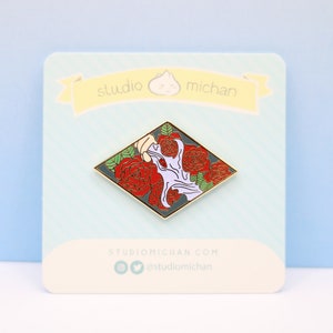 May include: A diamond-shaped enamel pin with a hand reaching into a bouquet of red roses. The hand is white with a purple outline. The background is dark gray. The pin has a gold border. The text "studio michian" is on the card behind the pin.
