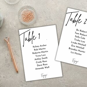 May include: Two white table cards with black text. "Table 1" and "Table 2" are written in a cursive font. Each card lists names. The word "Enjoy!" is at the bottom of each card. A rose gold pen and other stationery items are in the background.