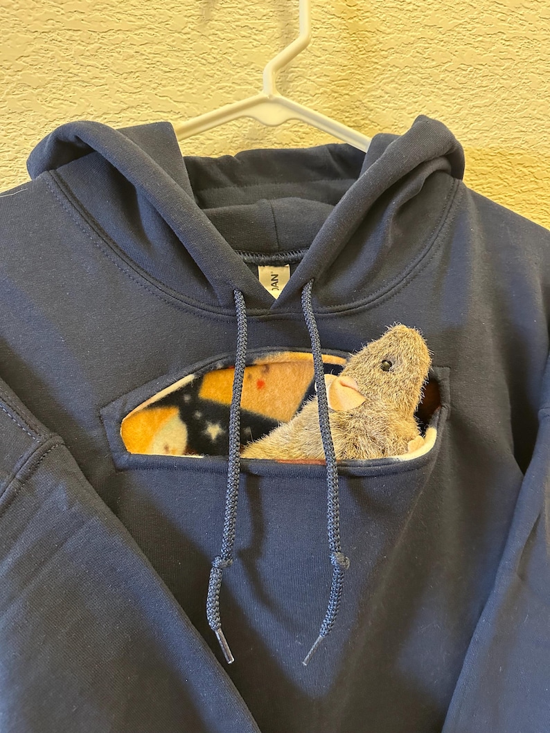 May include: A navy blue hoodie with a pocket on the front. The pocket has a fleece lining and a small stuffed animal peeking out of it.