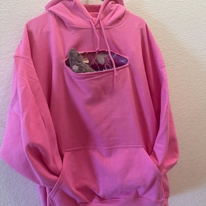 May include: A pink hoodie with a small pocket on the front. The pocket has a hole cut out of it, and a small stuffed animal is peeking out of the hole.