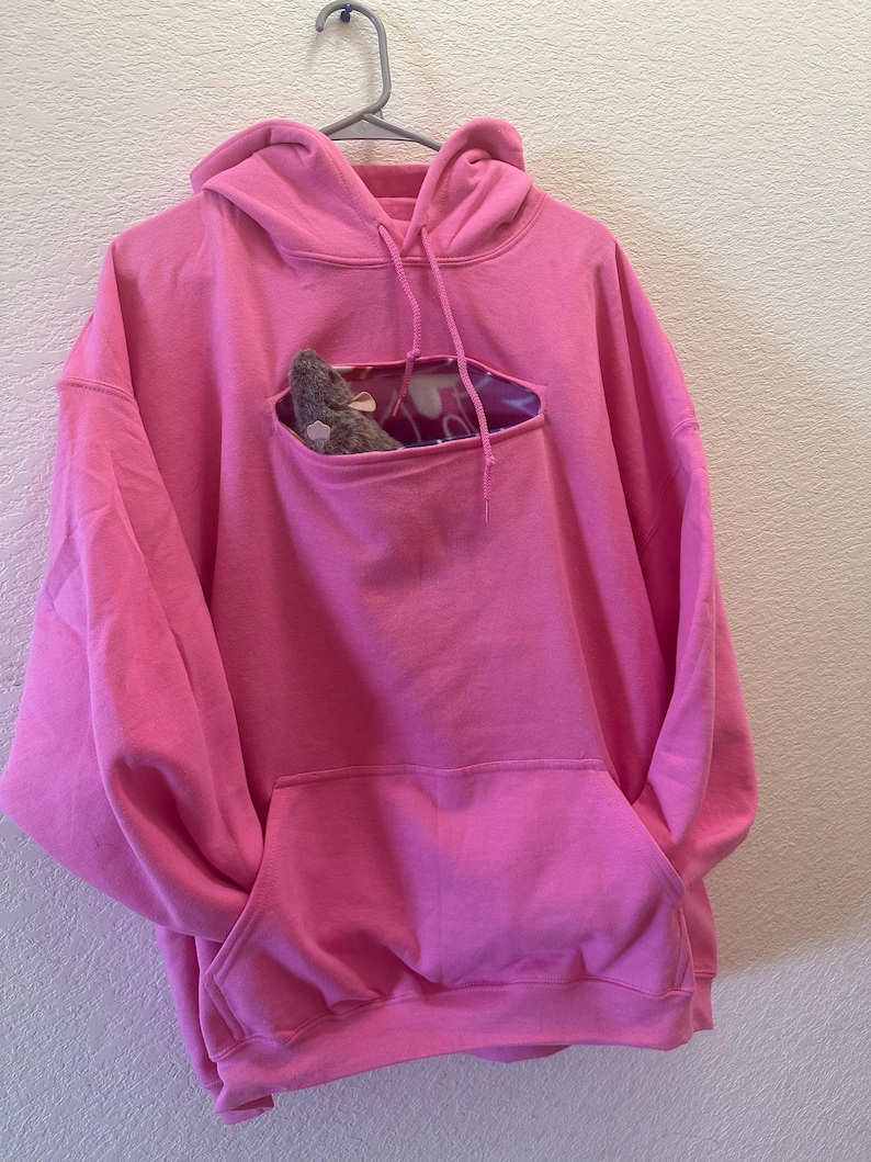 May include: A pink hoodie with a small pocket on the front. The pocket has a hole cut out of it, and a small stuffed animal is peeking out of the hole.