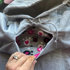 May include: A grey hooded sweatshirt with a fleece lining featuring a paw print and heart pattern. The hood is pulled back to reveal the lining.
