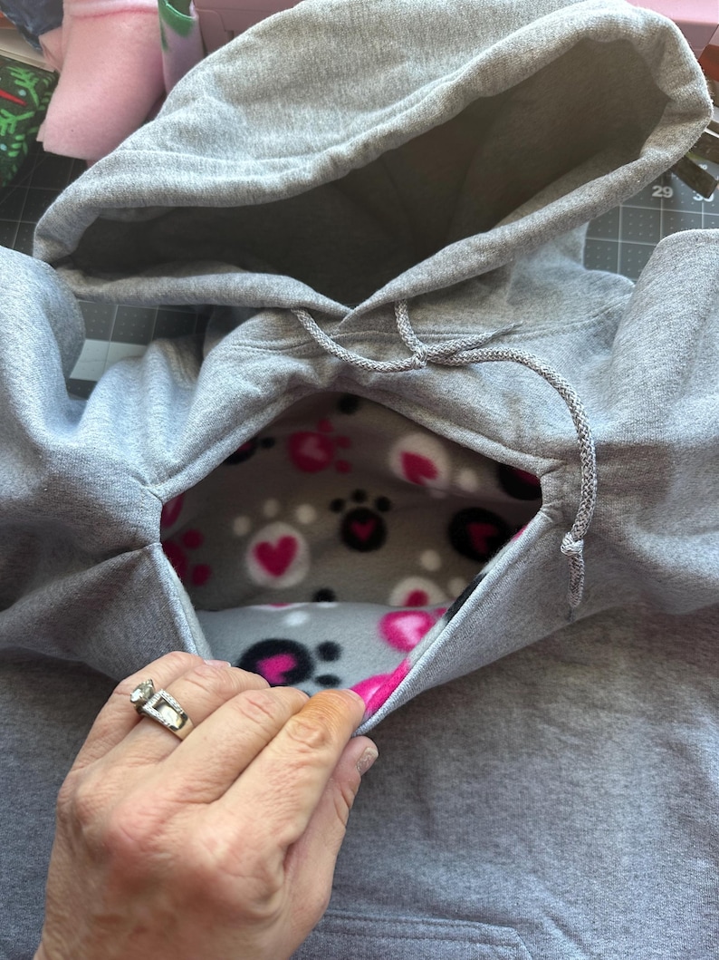 May include: A grey hooded sweatshirt with a fleece lining featuring a paw print and heart pattern. The hood is pulled back to reveal the lining.