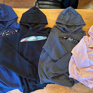May include: Four hooded sweatshirts in different colours: navy blue, black, grey, and pink. Each sweatshirt has a unique design on the front, with a cut-out revealing a patterned fabric underneath.