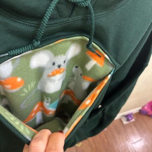 May include: A dark green hoodie with a hidden pocket lined with a green, white and orange fabric featuring a repeating pattern of mushrooms and white animals. The pocket is open and the lining is visible.