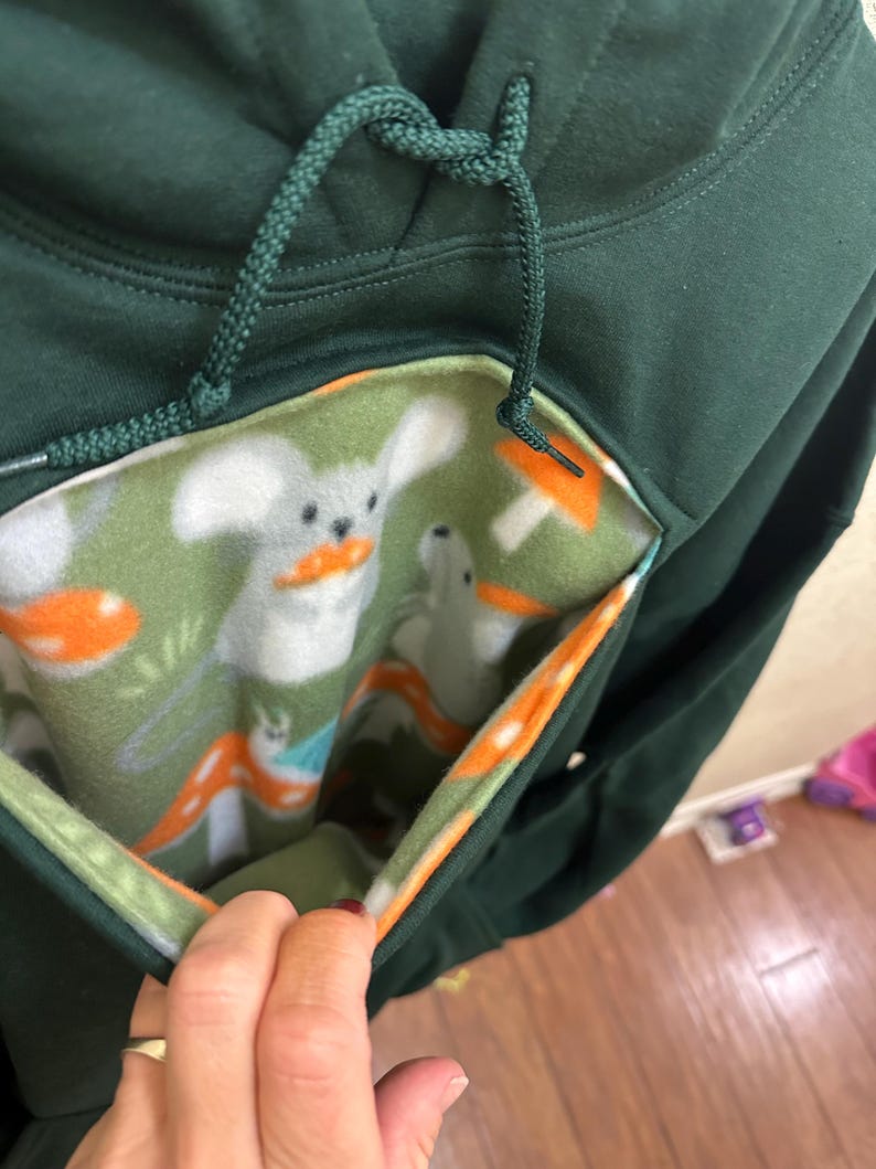 May include: A dark green hoodie with a hidden pocket lined with a green, white and orange fabric featuring a repeating pattern of mushrooms and white animals. The pocket is open and the lining is visible.