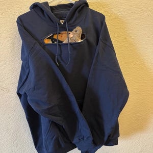 May include: A navy blue hoodie with a pocket on the front. The pocket has a cut-out shape that reveals a plush toy rat inside.