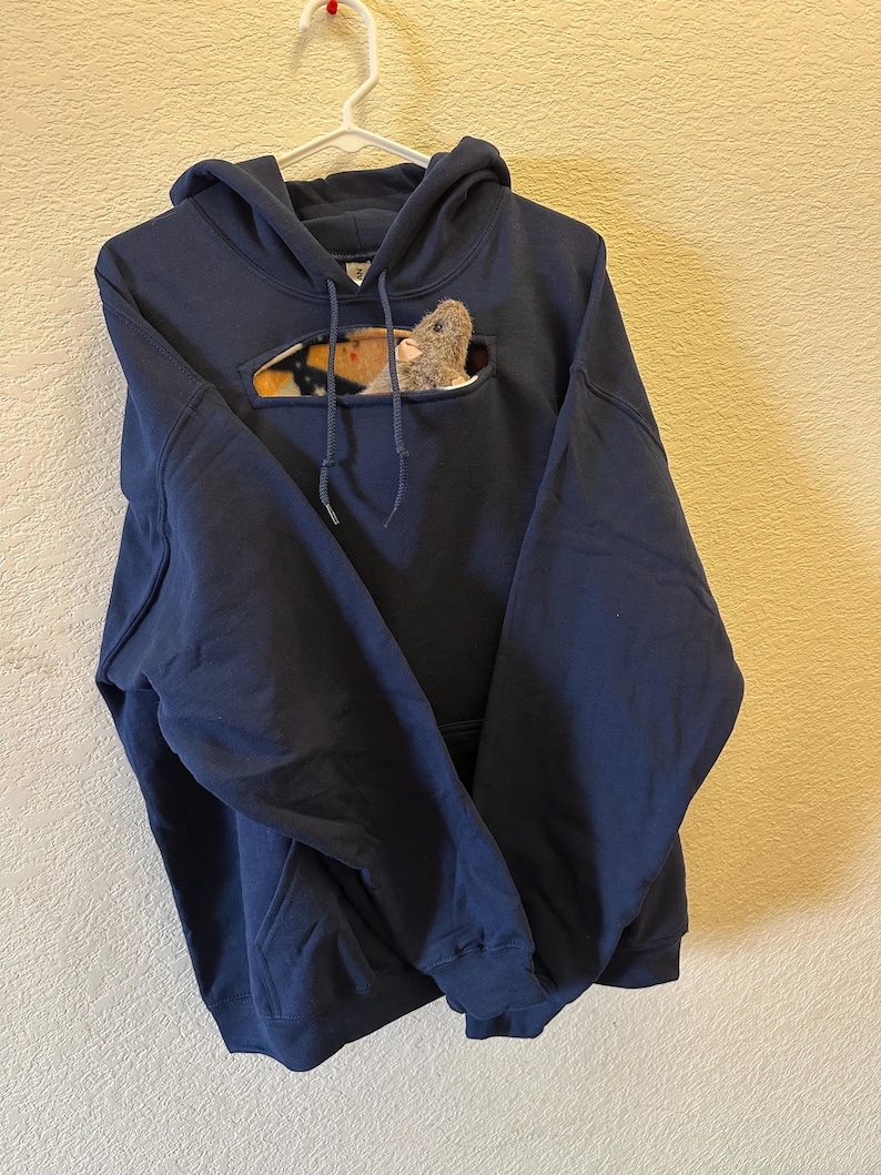 May include: A navy blue hoodie with a pocket on the front. The pocket has a cut-out shape that reveals a plush toy rat inside.