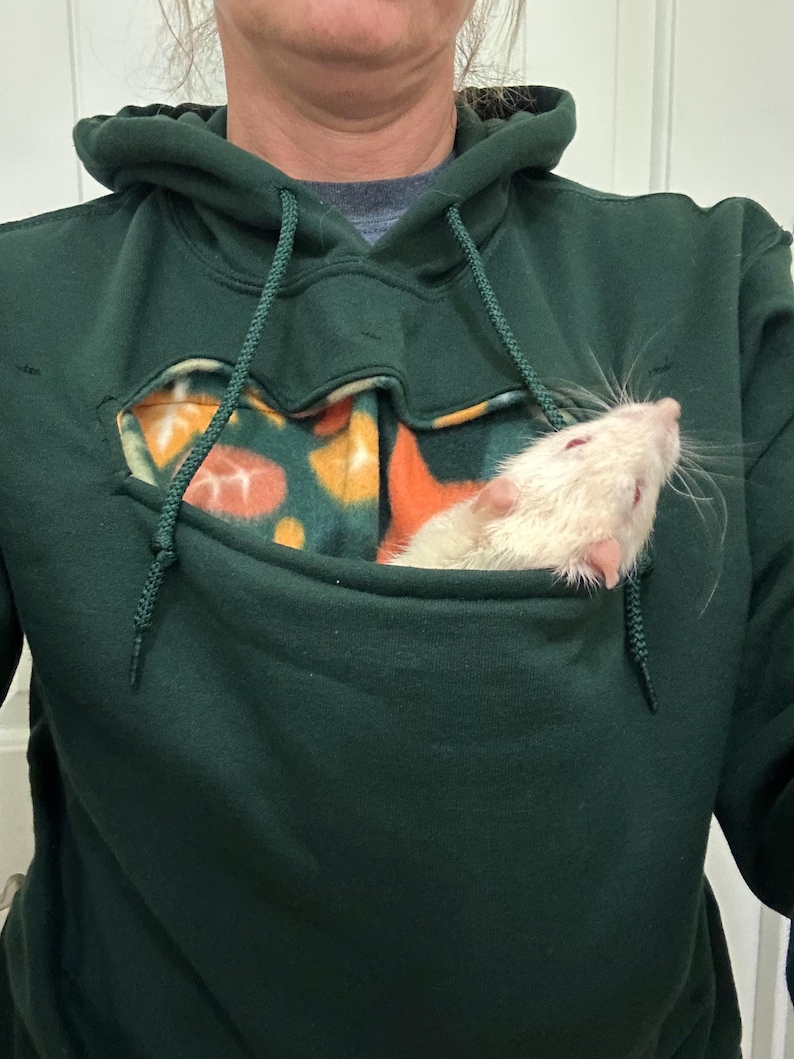 May include: A dark green hoodie with a fleece-lined pocket. A white rat is sitting in the pocket.