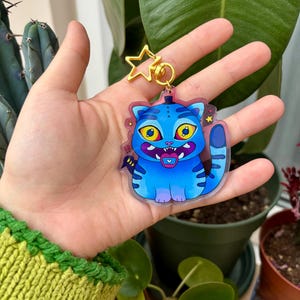 May include: An acrylic keychain featuring a cartoon cat with blue fur, dark blue stripes, and large yellow eyes. The cat has a wide grin with visible teeth and a small heart on its tongue. It is attached to a gold-colored star-shaped metal clasp.