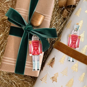 May include: Two wrapped gifts with a nutcracker and a bell ornament. One gift is wrapped in brown paper with a green velvet ribbon. The other gift is wrapped in white paper with a gold tree pattern and a brown velvet ribbon.
