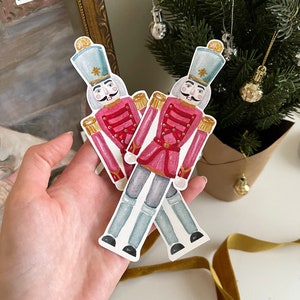 Puede incluir: Dos adornos de cascanueces de acuarela con chaquetas rojas, botones dorados y sombreros azules. Los adornos están sobre un fondo blanco.