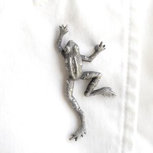 Puede incluir: Figura de rana plateada con textura detallada, en pose de escalada. La rana tiene las extremidades extendidas y patas palmeadas. El fondo es blanco, resaltando el diseño intrincado de la rana. La figura es de metal.