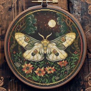 May include: Counted cross-stitch pattern of a white moth with brown and yellow accents, surrounded by green foliage and red and yellow flowers, against a dark blue background with a full moon and stars. The pattern is in a round wooden embroidery hoop.