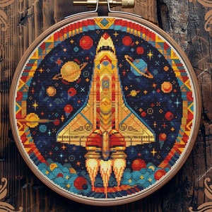 Op de afbeelding: Een geteld kruissteekpatroon met een ruimteschip in een kleurrijke galaxie met sterren, planeten en een blauwe nevel. Het patroon is ingelijst in een ronde houten ring. De tekst "Counted cross-stitch pattern by StitchingLand ©" staat onderaan de afbeelding.