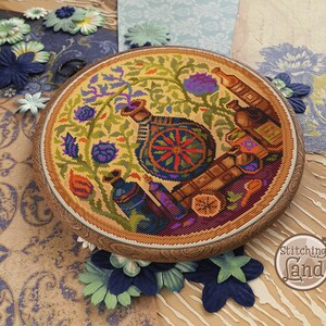 May include: A circular cross-stitch embroidery design featuring a colourful floral arrangement with a vase, bottles, and a wheel. The design is framed in a wooden hoop and has a yellow background.