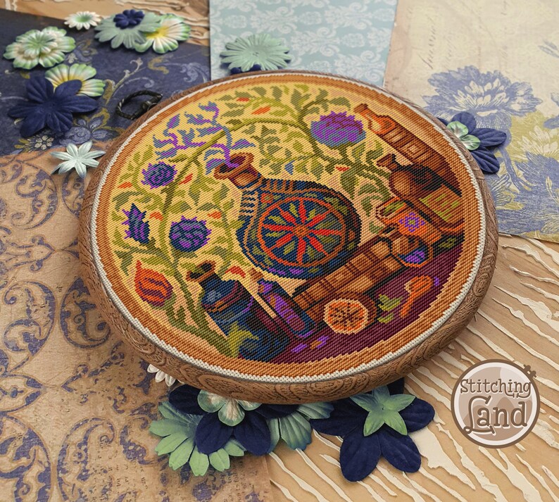 May include: A circular cross-stitch embroidery design featuring a colourful floral arrangement with a vase, bottles, and a wheel. The design is framed in a wooden hoop and has a yellow background.