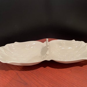 May include: A white, divided serving dish with a decorative, scalloped edge. The dish has two separate compartments and a handle in the center. The dish is made of ceramic and is suitable for serving appetizers or snacks.