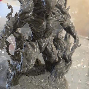 May include: A grey, 3D-printed figurine of a muscular humanoid figure with a textured surface. The figure is surrounded by stylized flames and stands on a cracked, circular base. The design suggests a fantasy or sci-fi theme.
