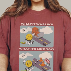 Puede incluir: Una camiseta roja con un diseño gráfico de una cara sonriente y un camino. El texto "What it was like" y "What it's like now" está impreso en la camiseta.