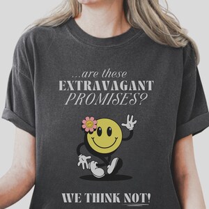 May include: A dark gray t-shirt with a white graphic of a smiling face with a flower on its head. The text on the shirt reads "...are these EXTRAVAGANT PROMISES? WE THINK NOT!"