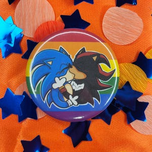 May include: A round button featuring a cartoon image of Sonic the Hedgehog and Shadow the Hedgehog kissing, set against a rainbow background. The button is surrounded by orange, blue, and pink confetti.