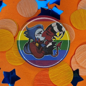 May include: A round button with a cartoon illustration of two characters embracing, set against a rainbow background. The button is surrounded by orange, blue, and wooden confetti.