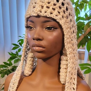 Könnte beinhalten: Ein beigefarbener Häkelhut mit langen, fließenden Strähnen. Der Hut hat ein einzigartiges Design mit einem quadratischen Muster. Der Hut befindet sich auf einer Puppe mit brauner Haut.