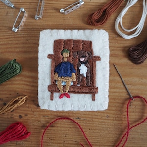 May include: A felt embroidery of a person and a dog sitting on a bench. The person is wearing a blue shirt and a brown hat. The dog is brown and white. The embroidery is on a white background.